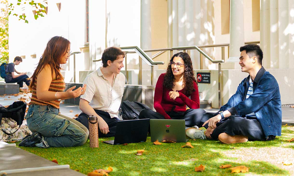 Four students sitting and talking outdoors on campus, using laptops, surrounded by autumn leaves and sunlight.
