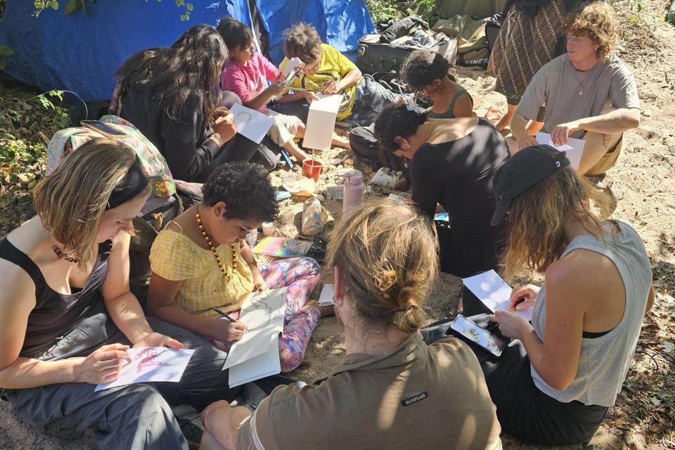 A group of people sitting outdoors on the ground, sketching or writing in notebooks. 