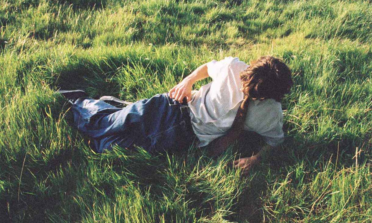 A person in jeans, a white short-sleeved shirt and a brown tie lounges in long green grass.