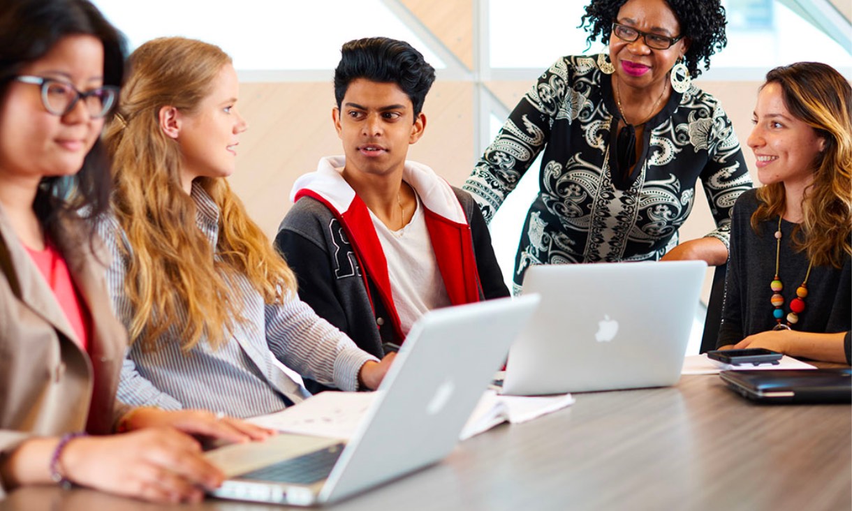 An older female teacher interacting with high school students in a classroom.