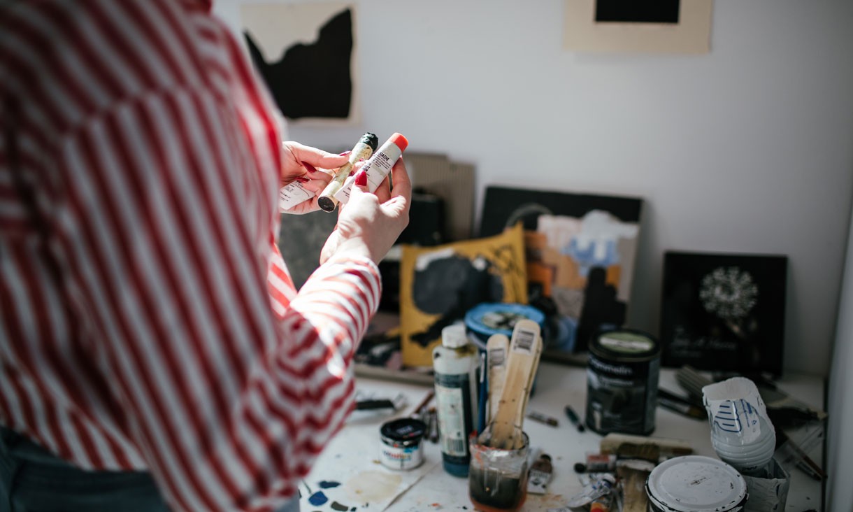 A person holds a pair of black and red paint tubes in front of her work station.