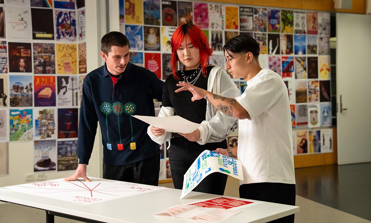 Three people discuss and review design posters on a table in a creative classroom space filled with colourful artwork on the walls.