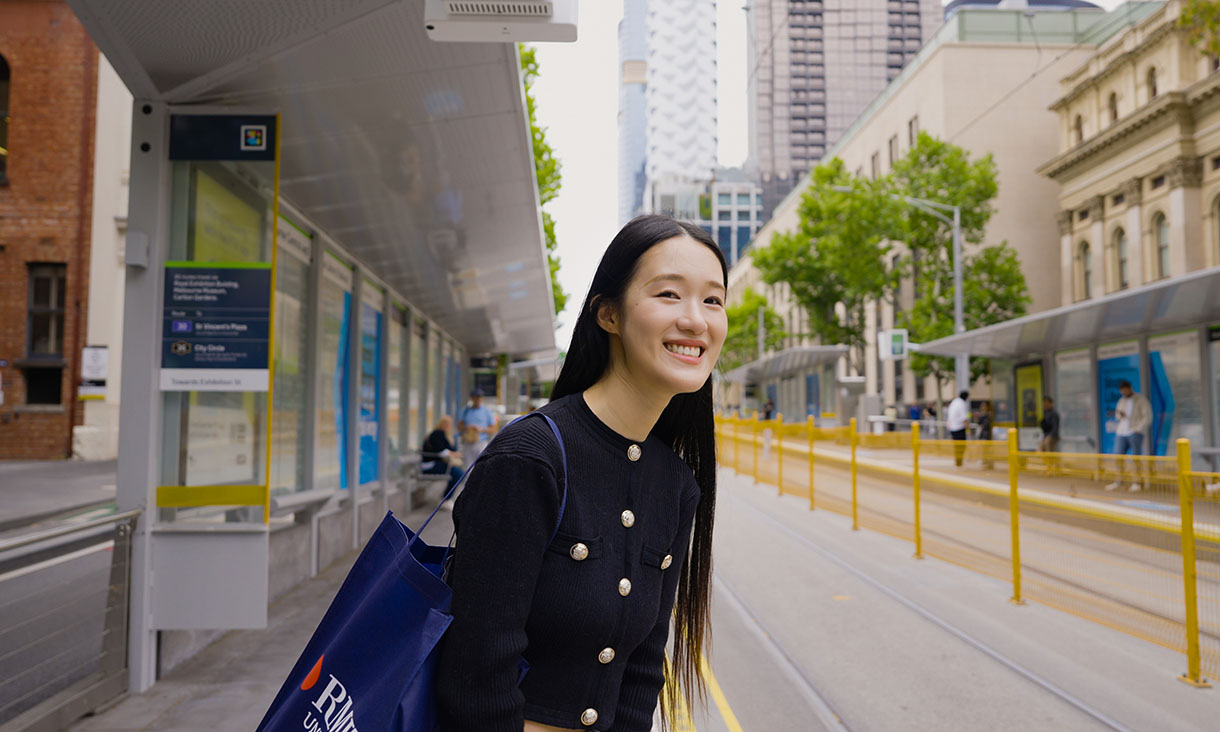 RMIT student smiling while waiting at a tram stop as a tram approaches.