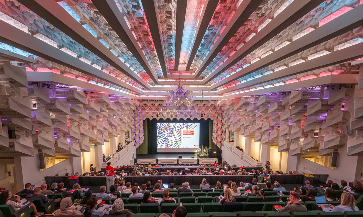 Audience seated in a large lecture theatre facing a presentation screen, beneath a dramatic ceiling with pink, blue and white lighting.