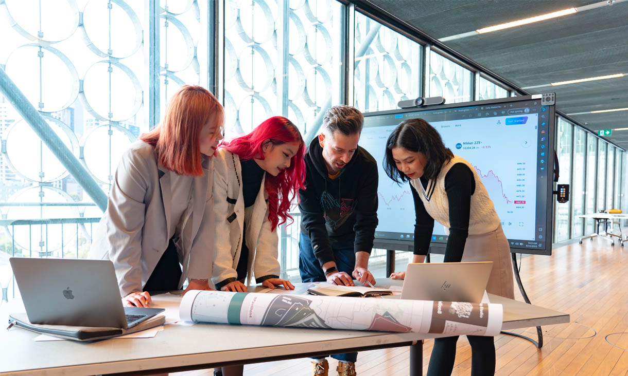 RMIT Business students stand about a table comparing notes