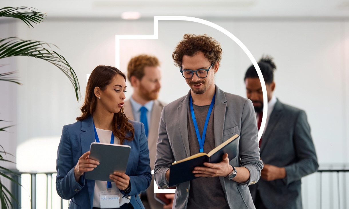 Two people conversing in a corporate environment, comparing notes using a tablet and a notebook.