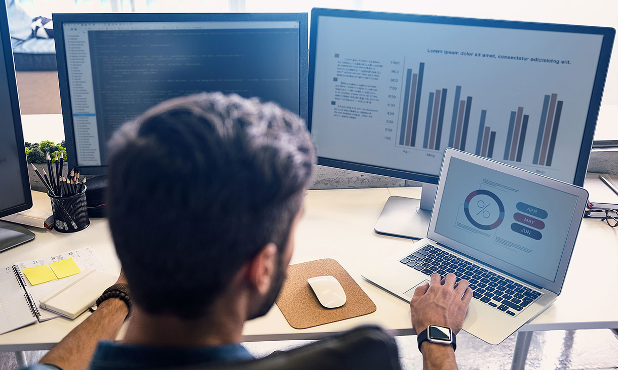 Man works on laptop and two screens facing away from camera