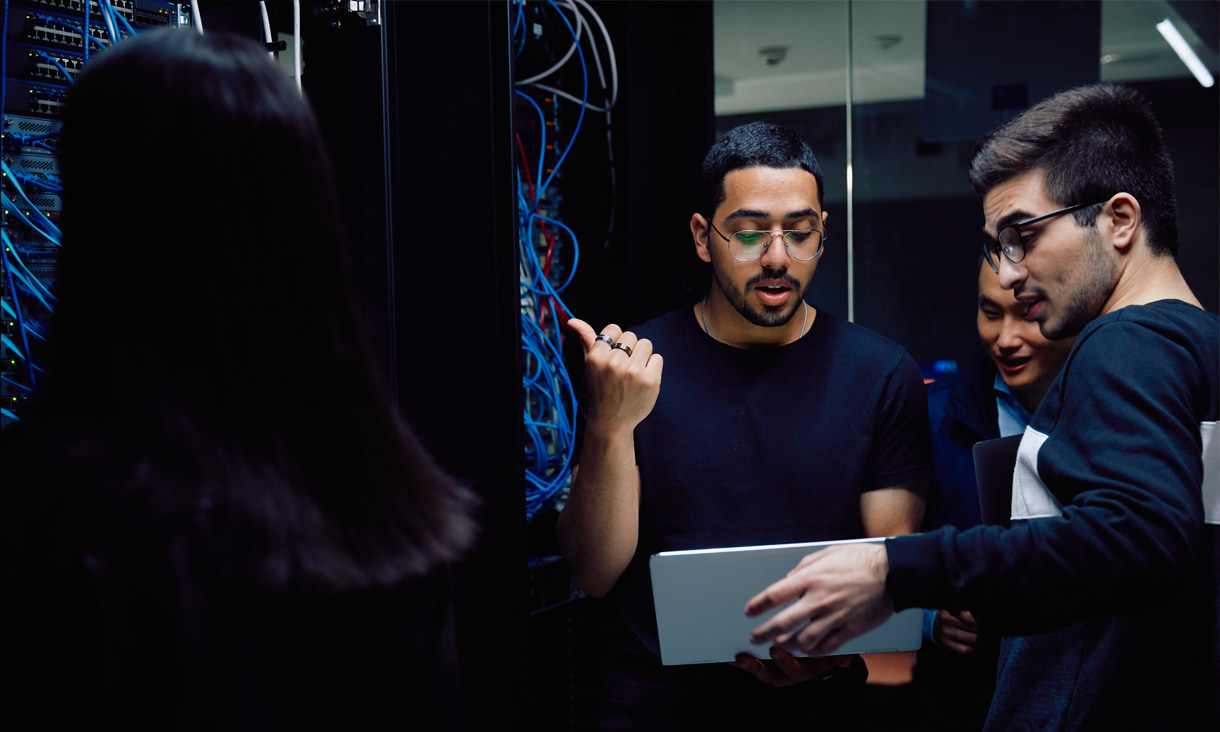 Three young people standing with a laptop next to a server bustling with cables
