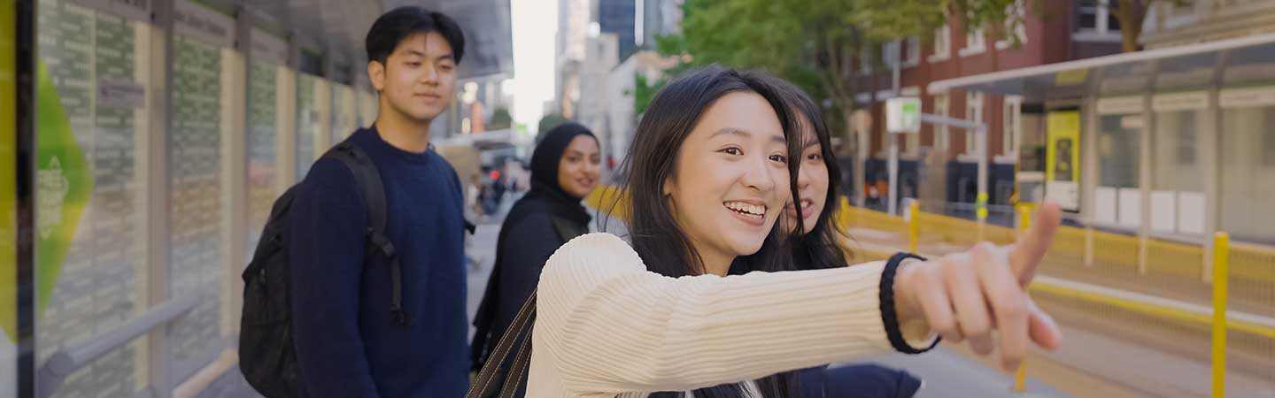 Four smiling students at a tram stop, one pointing.