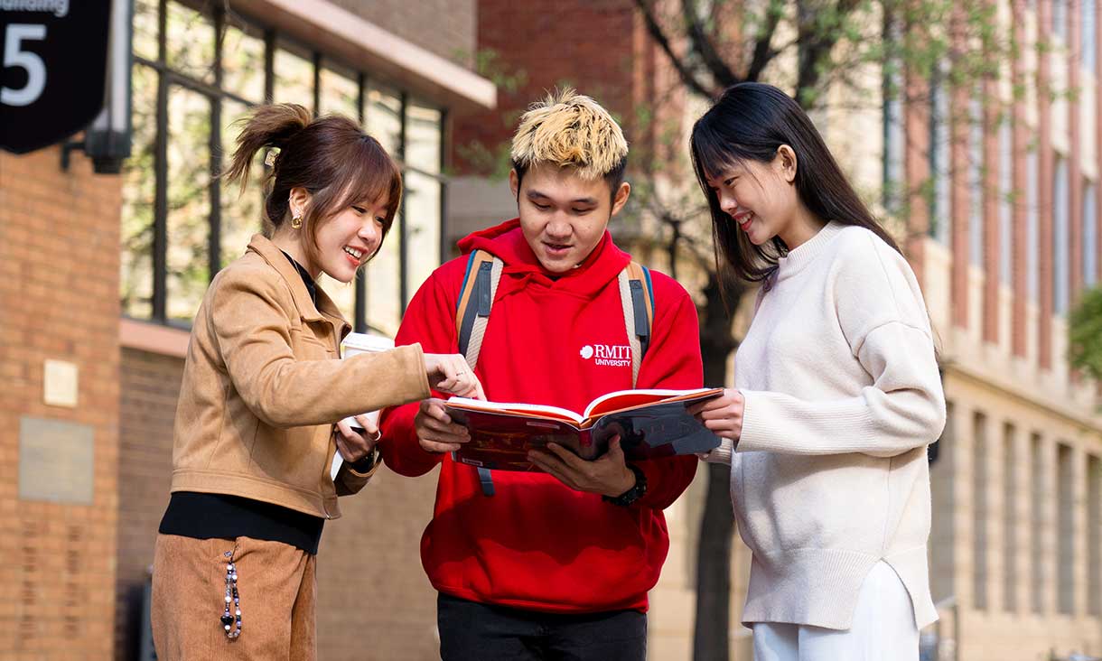 Three students standing in the Melbourne city campus and looking at a text book.