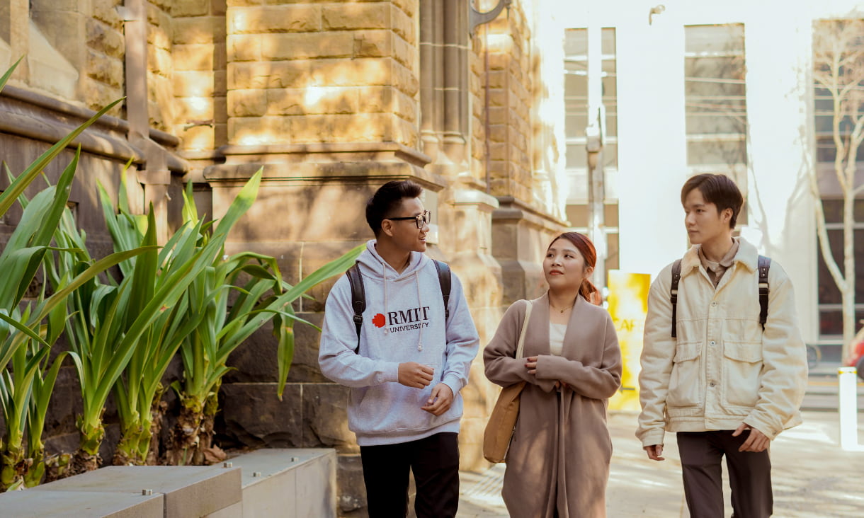 Three students with backpacks walking and talking together outside a historic building at RMIT University on a sunny day.
