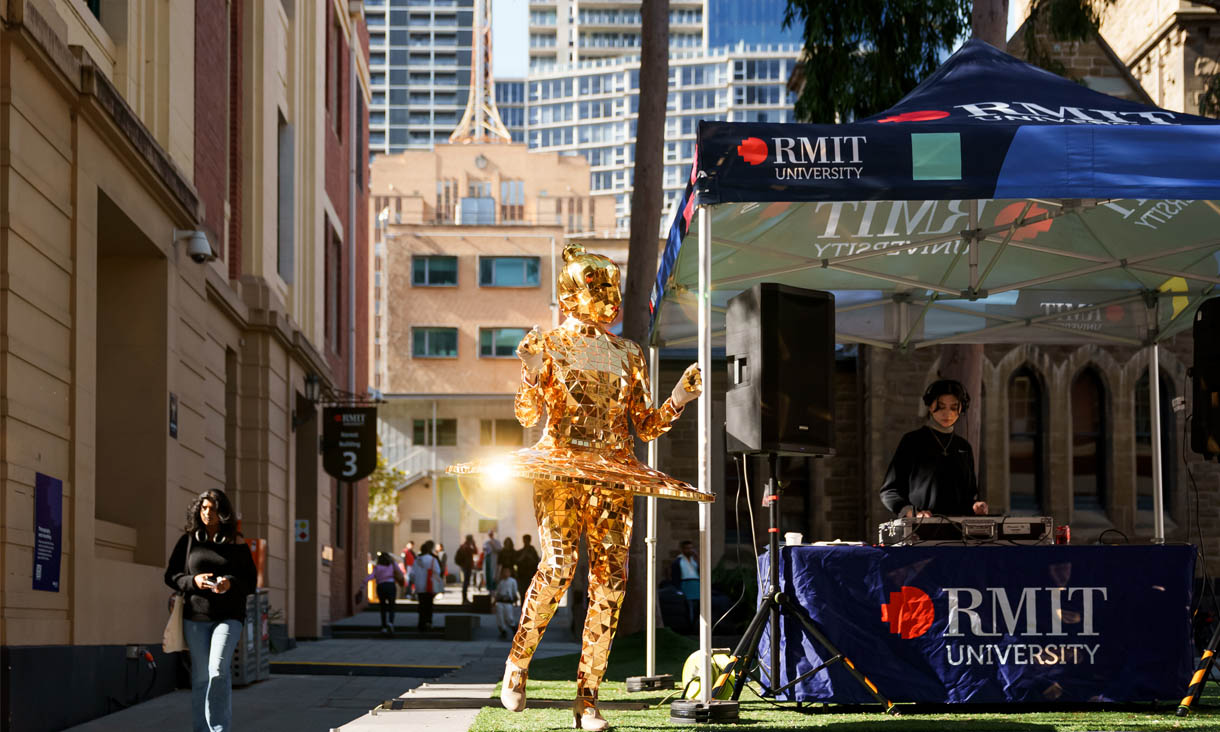 A performer in a golden mosaic costume dancing beside a DJ in an RMIT branded marquee