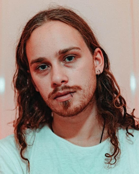 Headshot of Hayden Ryan on a dark background. Light halos the top of his head. He has long, dark, wavy hair and facial hair and lip piercings.