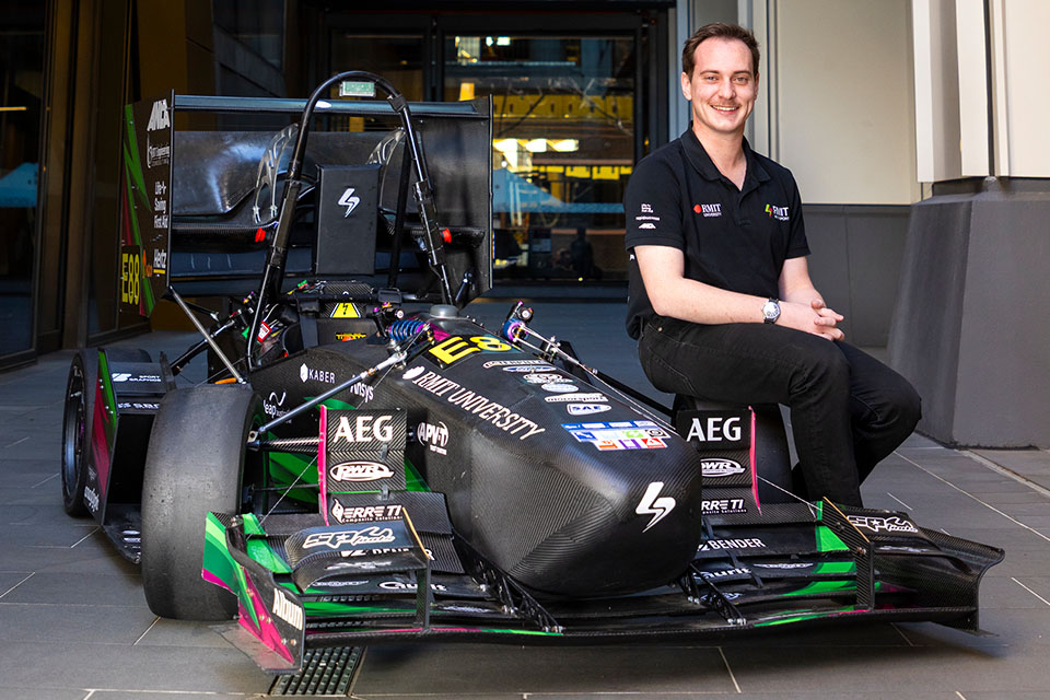 A person in an RMIT-branded team polo shirt sits beside a low, black Formula-style racing car covered in sponsor logos and the words 'RMIT University', parked in an outdoor campus courtyard.