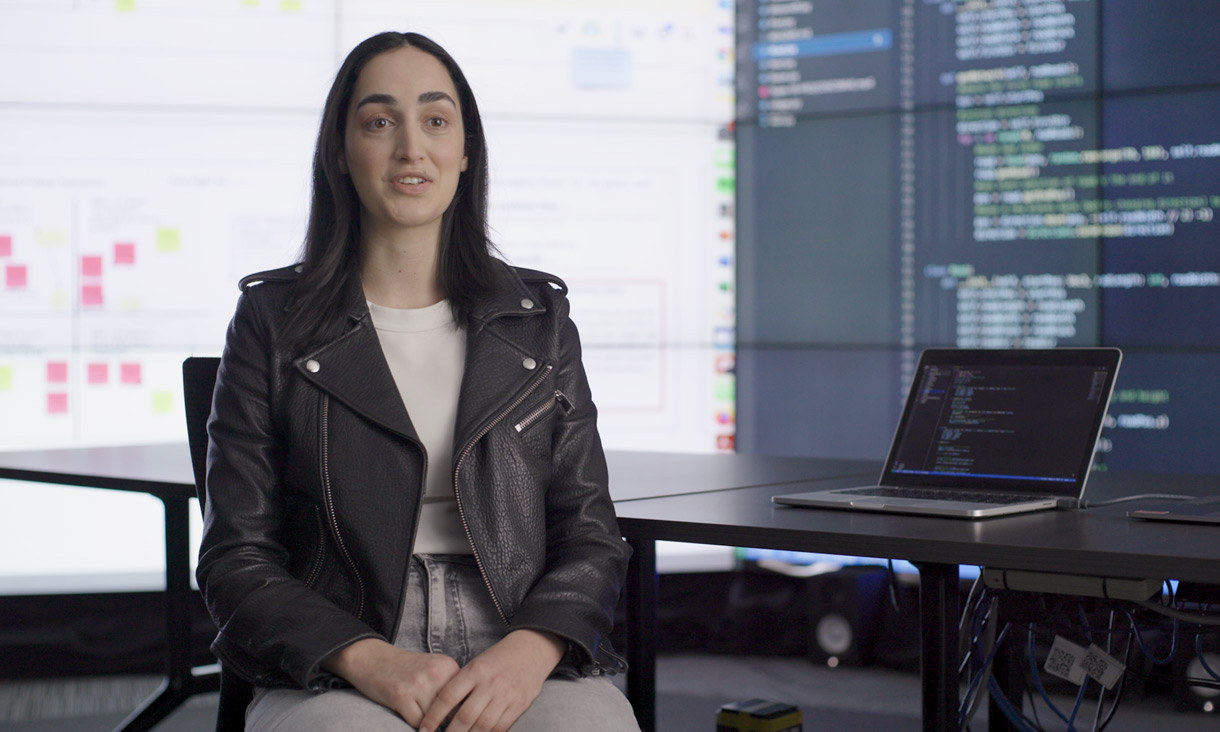 Nicole, talking to camera, in front of large screens filled with computer code