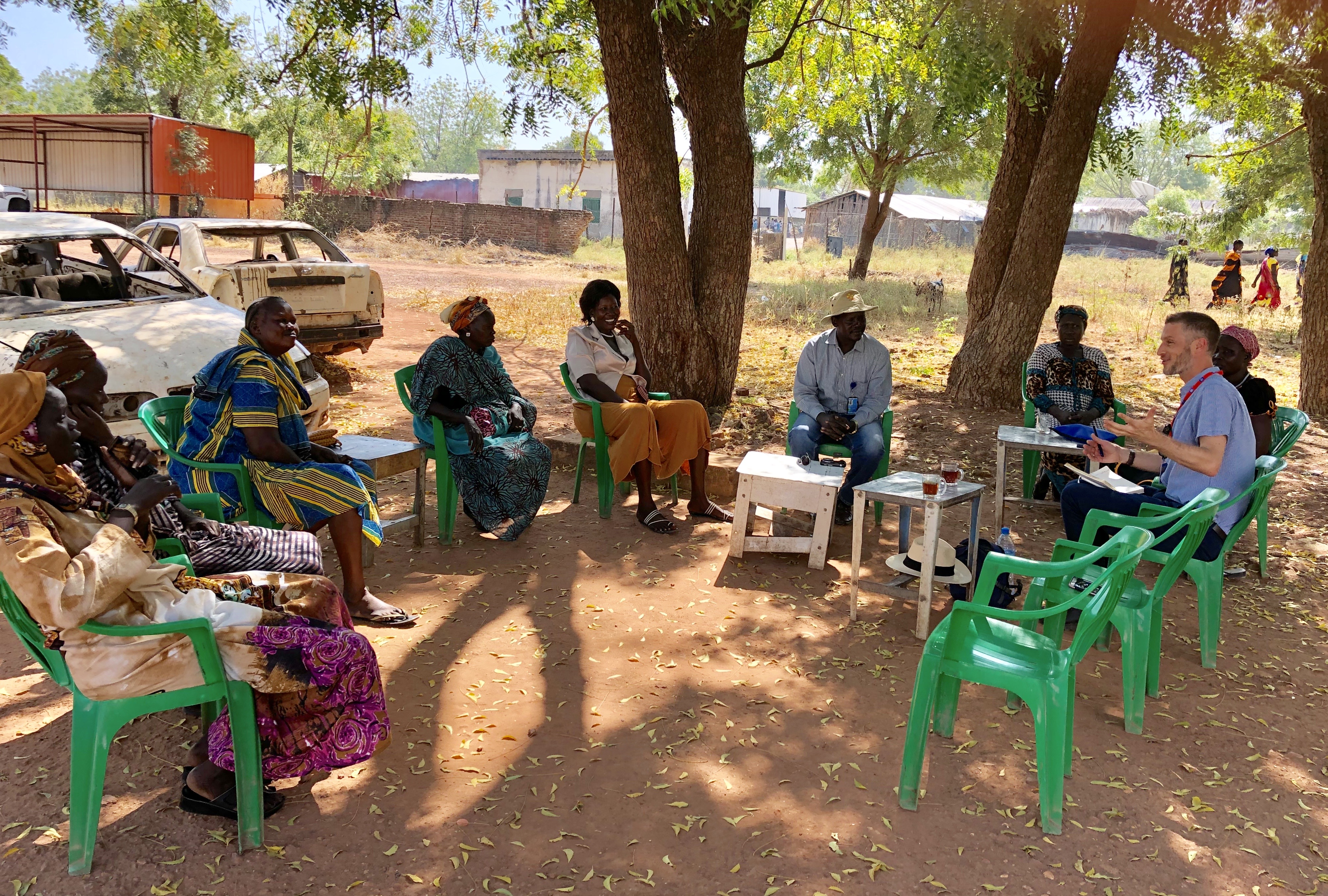 Photo of a group of people sitting outside. Cars are near the group, to the left of the photo. The group is shaded by two tall trees. The group are sitting on green plastic chairs. In the background are fences and buildings. The terrain is dusty and red, with yellow grass growing intermittently.