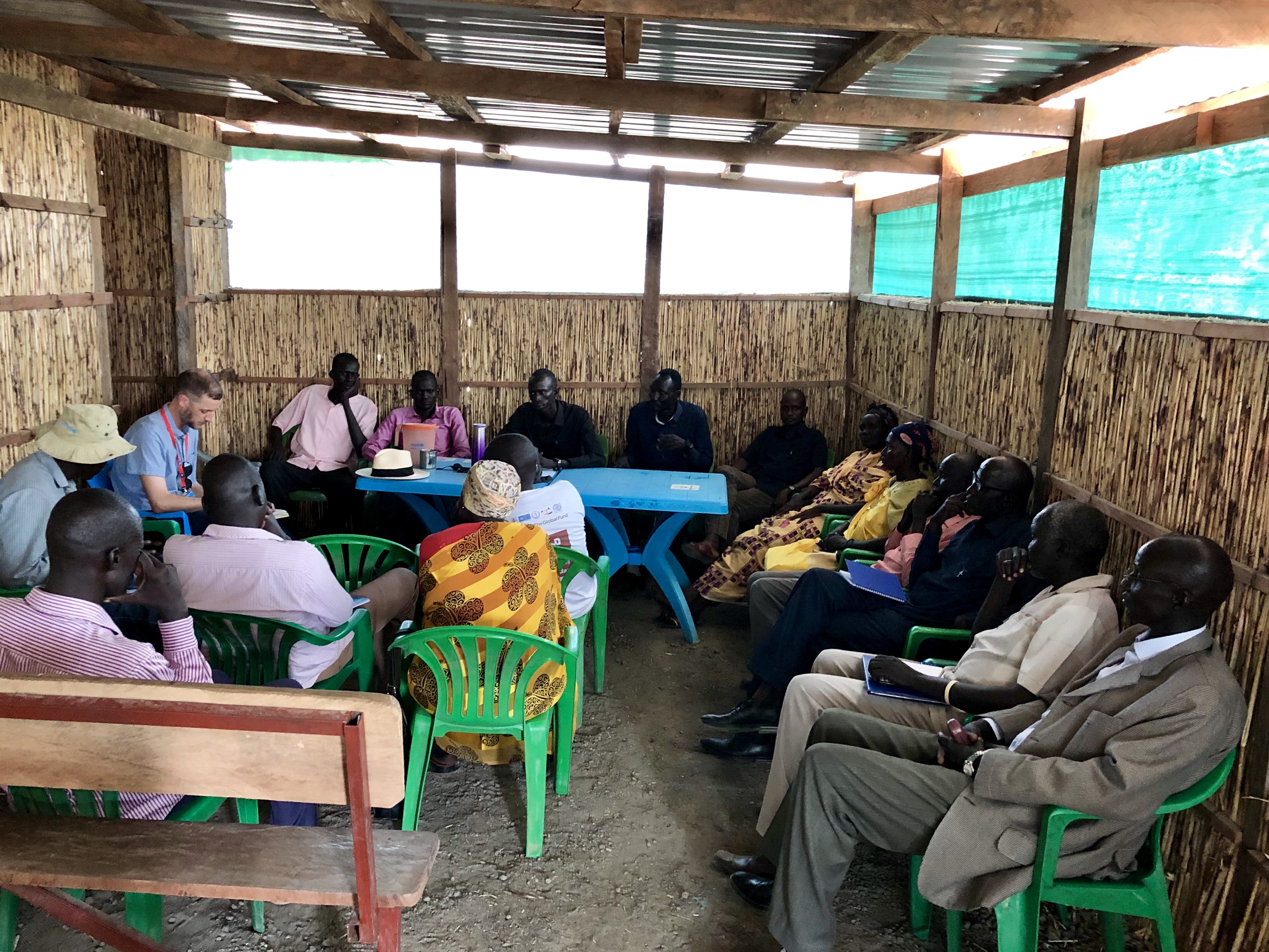 Photo of a group of people sitting inside. The surrounding walls, made of a natural material, go up half way. The rest is open to the outside. Above them is a tin roof. About 16 people are spread out, some sitting on wooden benches, others on plastic chairs.