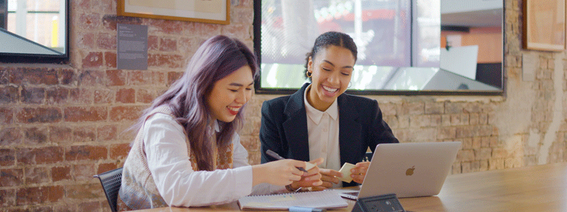Two smiling women seated with a laptop and papers, in front of a brick wall with artwork. 