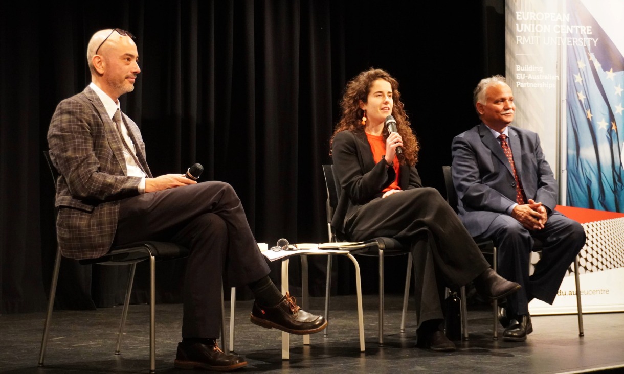 A woman sitting on a stage with a black curtain backdrop, speaking into a microphone. Two men in suits are also sitting in the panel, one holding a microphone.