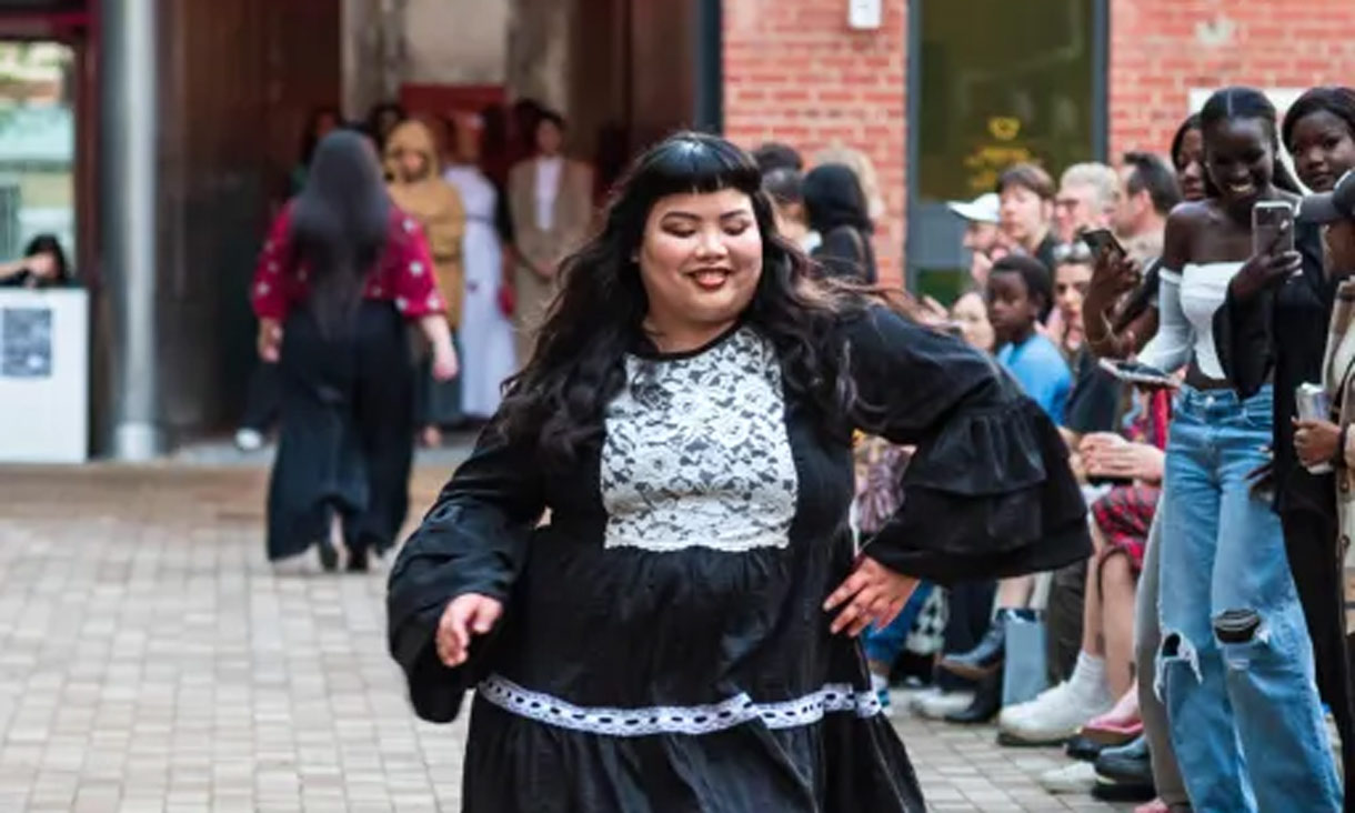 Person in a black dress with white lace detailing, walking forwards. On the right hand side, a group of standing people taking photos with mobile phones of the main person.