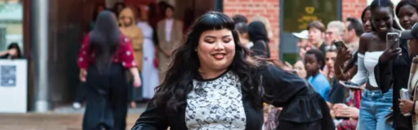 Person in a black dress with white lace detailing, walking forwards. On the right hand side, a group of standing people taking photos with mobile phones of the main person.