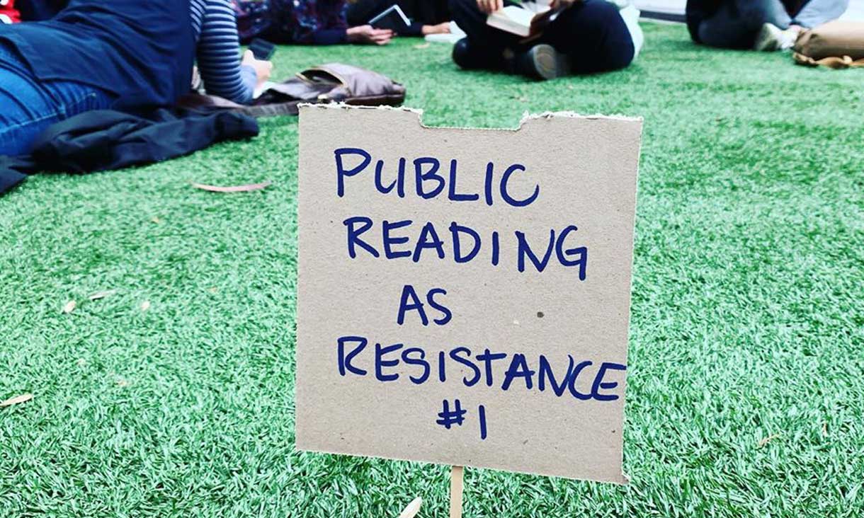 a photograph of people sitting outside on grass reading books. At the front of the image is a small handwritten sign on cardboard that says, 'PUBLIC READING AS RESISTANACE #1'.