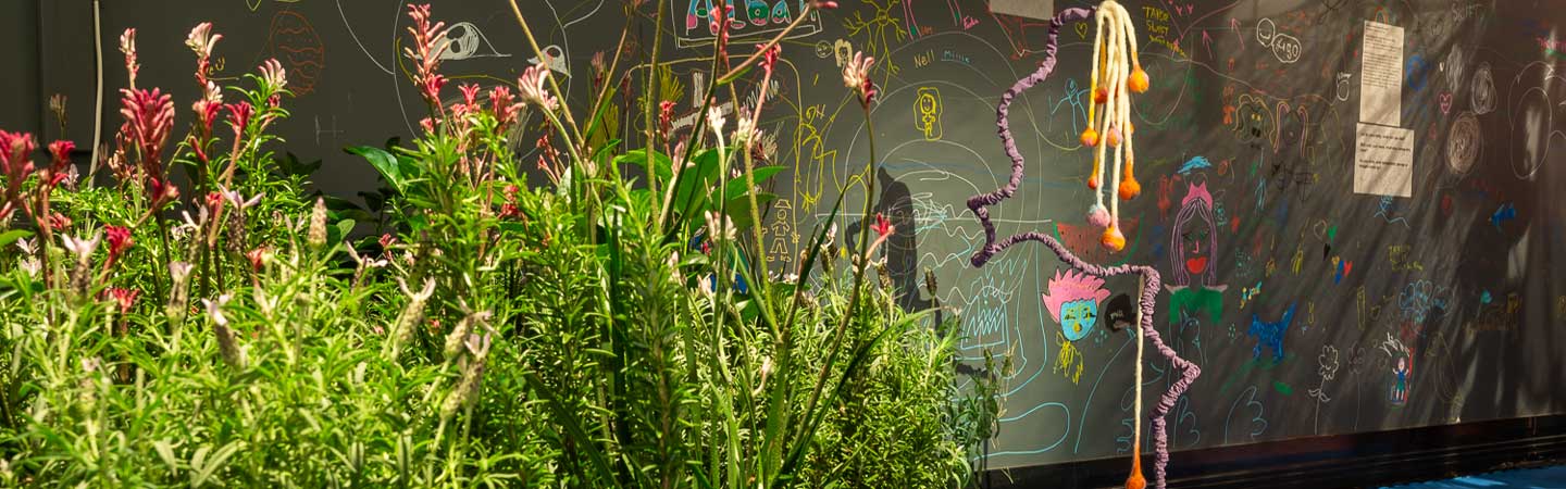 Bright green shrubs with pink flowers on a large timber platform, in the foreground. In the background, a chalkboard with children's drawings. There is also a thin and tall knitted sculpture on the floor which is blue.