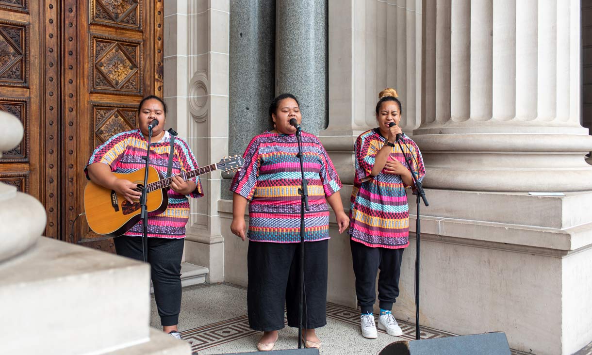 Three people stand in front of a building with light grey columns and a large brown wooden door. There are singing into the microphones which are held up by three stands. The person on the left is holding a guitar.