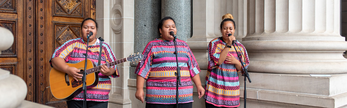 Three people stand in front of a building with light grey columns and a large brown wooden door. There are singing into the microphones which are held up by three stands. The person on the left is holding a guitar.
