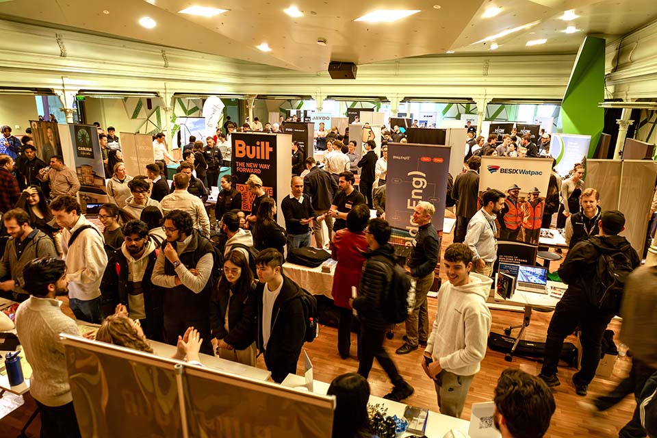 Crowded career expo hall with students visiting employer booths and banners, networking and exploring opportunities.