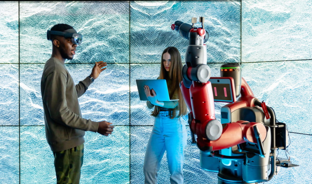 Two people from front of a large videoscreen. A man wears a VR headset and a woman holds a laptop.