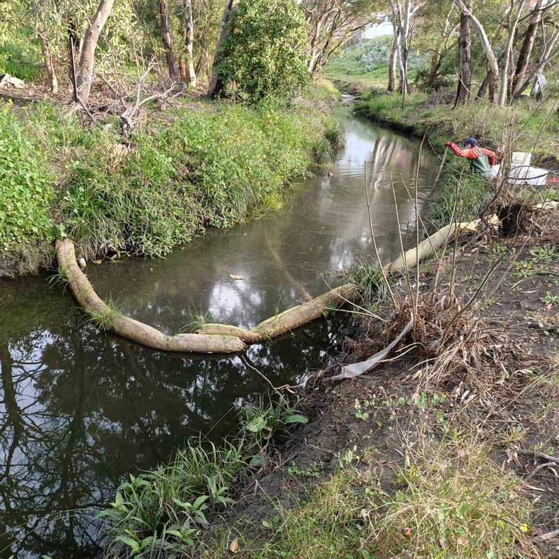  Photos of Stony Creek with a pollution barrier sausage. The creek is running from the bottom left to the top middle of the photo before being cut off by a hill. Towards the bottom of the photo is a brown-stained cylinder object running horizontally across the creek from one side to the other. Similar to a sausage link, the object is pinched in multiple areas. In the background is a researcher wearing a light red, long-sleeved top with green overalls and a dark blue hat. The researcher has some white storage containers behind them as is poking what looks like a long stick into the creek. The creek is surrounded by eroded dirt and green folliage, with native Australian trees.