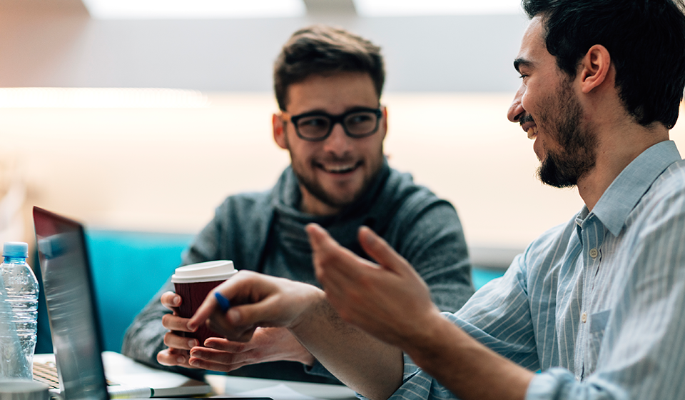 Two men having a converstion. One is holding a coffee cup. They are both smiling.