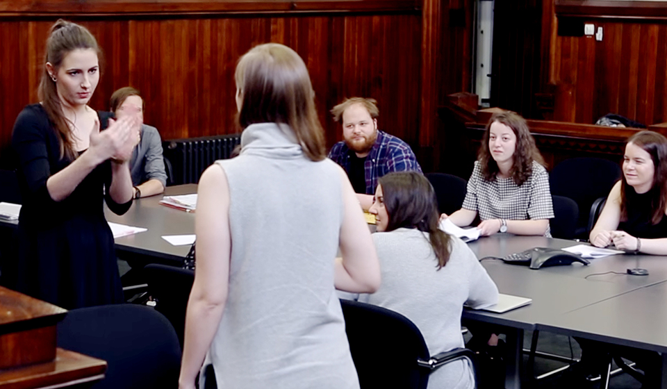 Female using hand motion to communication with other female student, while being looked at by seated students