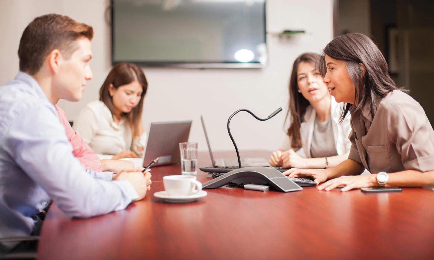 Four people seated at a large office meeting desk, talking into a microphone/telecom unit.