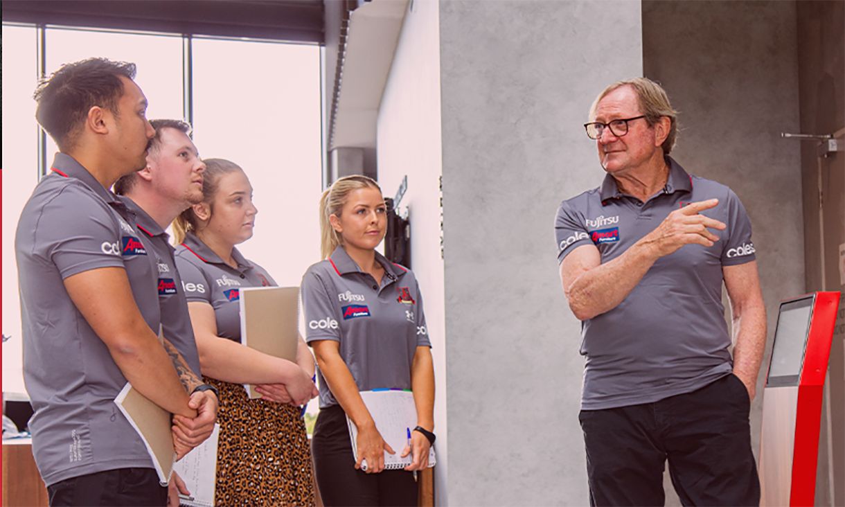 Male instructor speaking to four students in grey polo shirts.