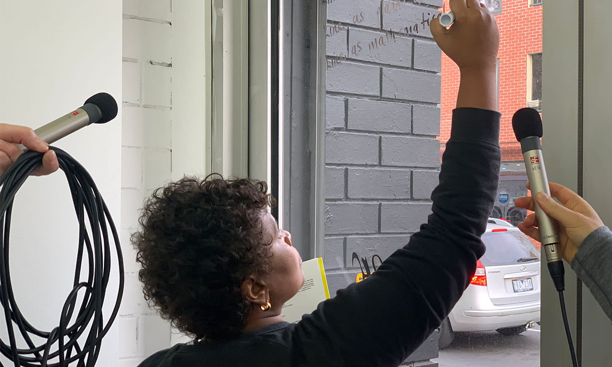 Amaara Raheem writes on a piece of glass. Two microphones are pointing towards her.