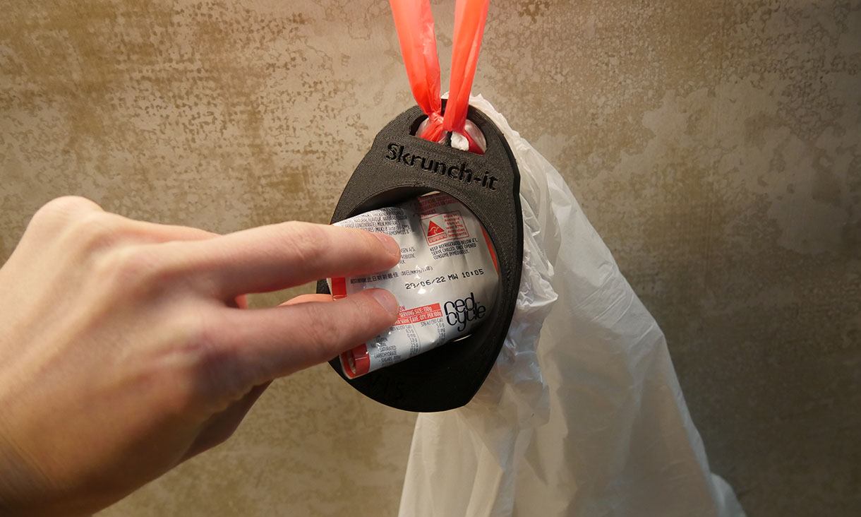 The Skrunch-It is attached to the top of a bin bag and someone is placing a piece of soft plastic inside.