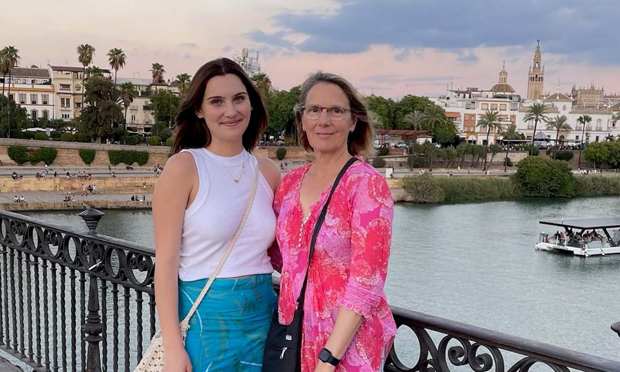 Young woman with brown hair and mature woman with grey hair stand on bridge over river smiling at camera