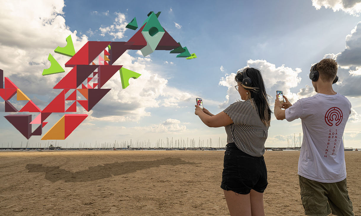 Two young people aim their phones at an abstract computer-generated image while standing on a beach.