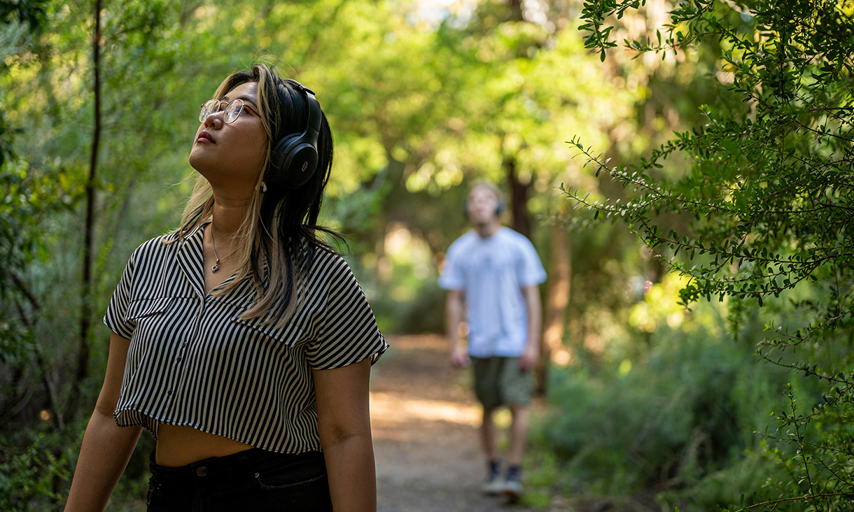 Someone wearing headphones wanders through natural bushland. A blurred figure wearing headphones is in the background also.