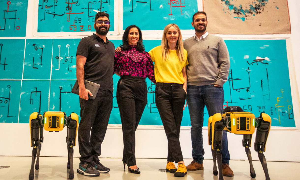 Four people stand with two robot dogs in front of a wall of art