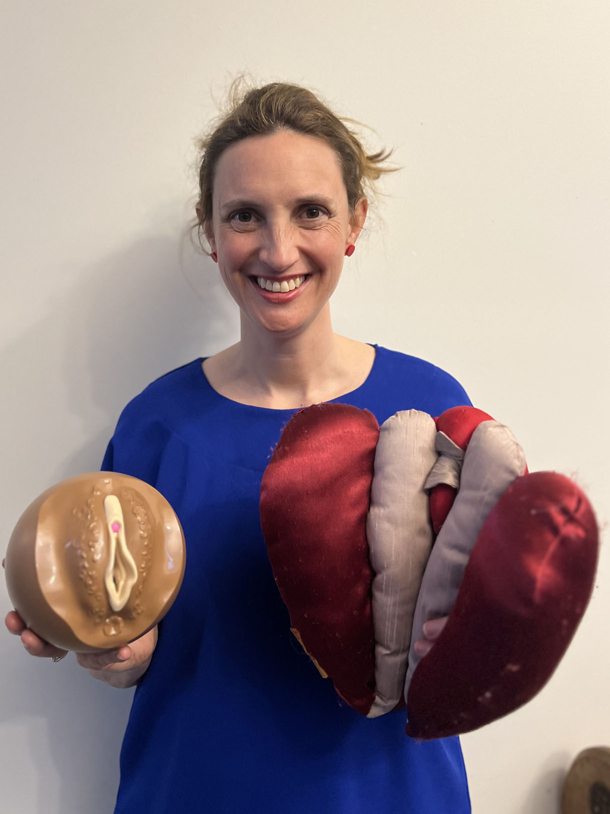 Anita Brown-Major stands smiling holding Cliterate, a pull apart model of the vulva, and a vulva puppet.
