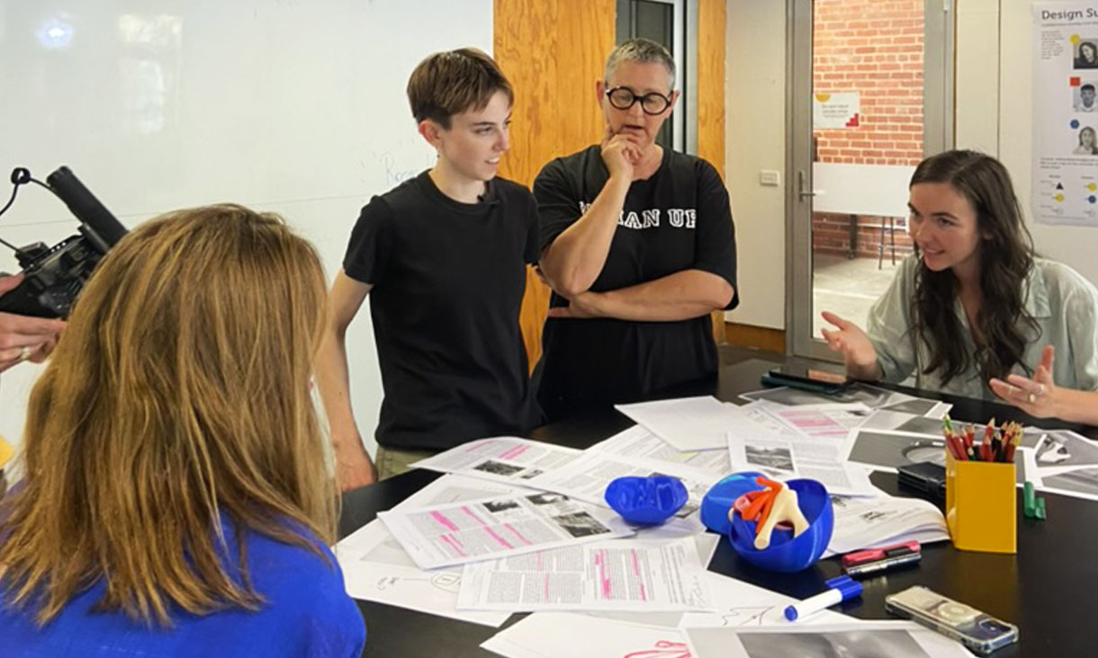 The design team behind Cliterate are standing around a table full of papers and a Cliterate prototype while discussing the project.