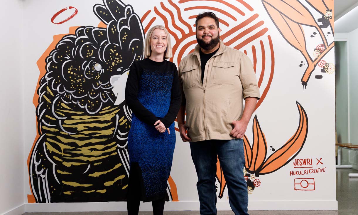Nicola St John and Emrhan Tjapanangka Sultan stand in front of a wall painting of a black cockatoo by Jeswri x Mukulri Creative.