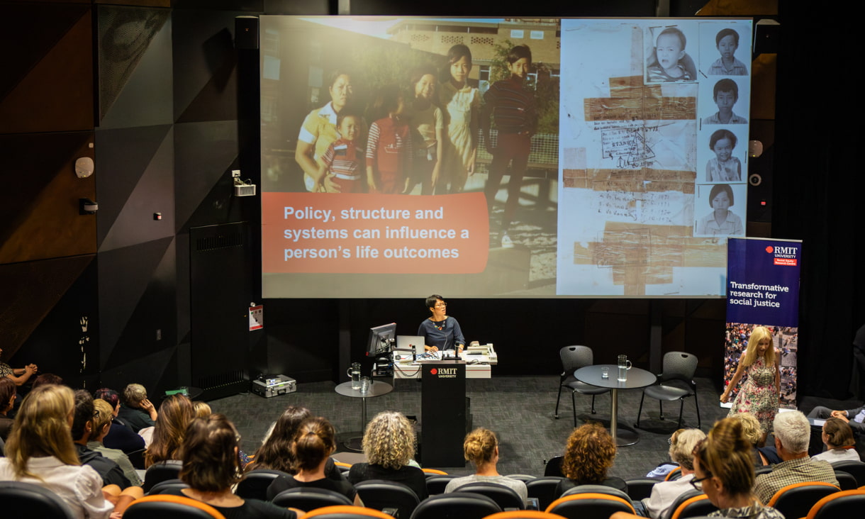A woman stand behind a podium with a projected image on a screen behind her and an audience in front of her.