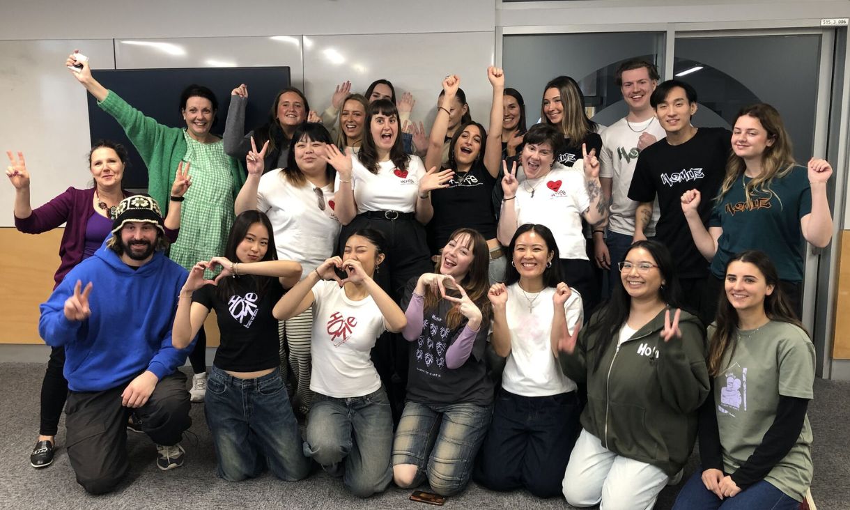 Students posing with teachers and HoMie co-founder, each with jubiliant expressions and hands in the air, celebrating the end of their pitches.