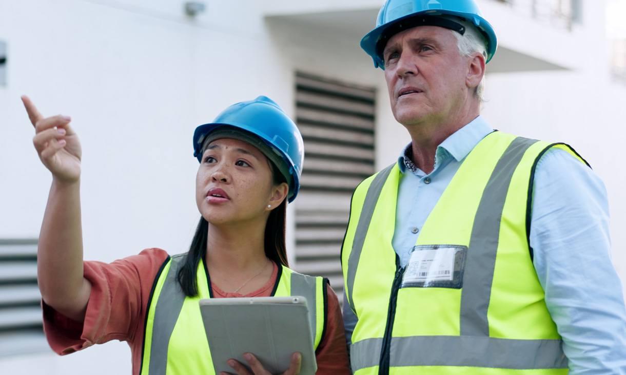 Two workers wearing hard hats and high vis vests at a construction site. One person is pointing at something in the distance. 