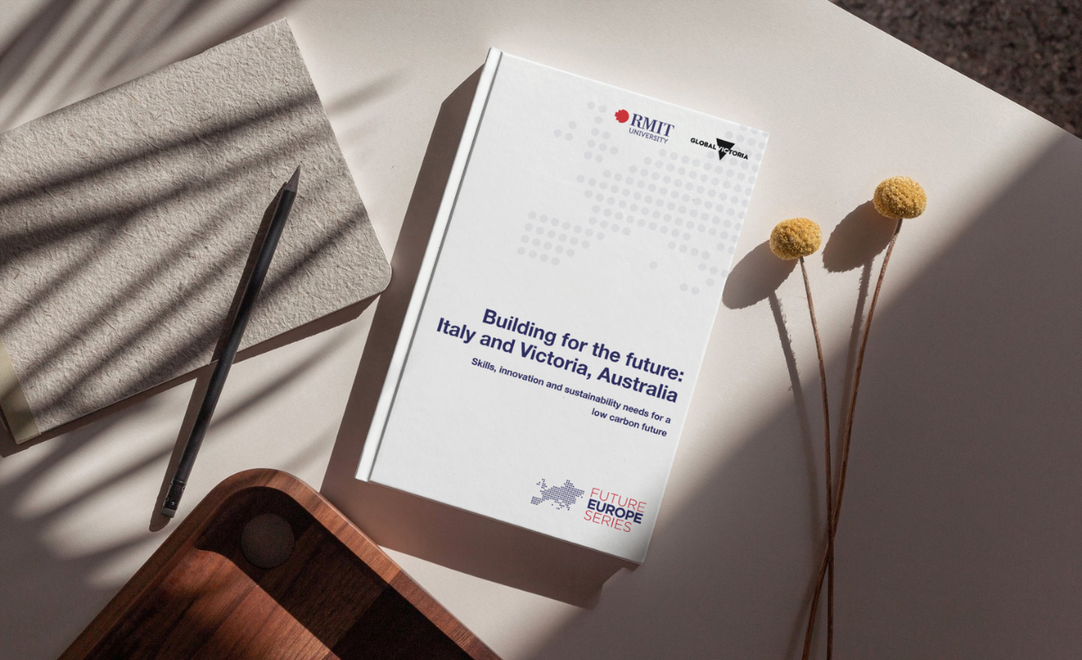 A book titled "Building for the future: Italy and Victoria, Australia" lies on a table with a notepad, pencil, and decorative flowers.