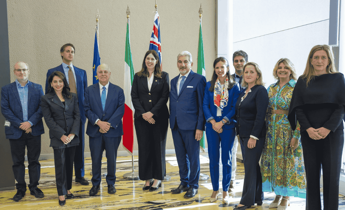 A group of people stands in front of European, Italian, and Australian flags in a formal setting.